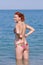 Ginger girl stands knee-deep in sea water and adjusts her bikini