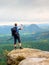 Ginger hair man takes photos with smart phone on rocky peak above autumnal valley.