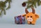 A ginger knitted bull stands next to a beautifully wrapped gift box. Behind him a cone hangs from a spruce branch.