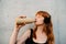 Ginger woman with headphones drinking water by concrete wall outdoors