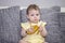 The girl of 10 months eats a ripe banana sitting on a gray sofa. A small child eats exotic fruits. Girl in yellow with a banana in