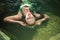 A girl with algae in her hair and in a green dress lies in the water on stones in a pond. Model with a rim with shells. Mermaid in