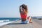 Girl with arms raised sits on seafront, rear view