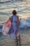 Girl from the back stands in the water on the beach, holds long dress upwards with hands