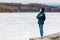 Girl with a backpack, young woman in a blue jacket and hood looks into distance at a frozen lake