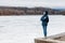 Girl with a backpack, young woman in a blue jacket and hood looks into the distance at a frozen lake