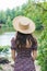 Girl backs stands by the lake in summer dress and straw hat.