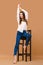Girl with bare feet and in casual outfit posing at tall wooden stool in studio