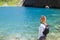 girl with black backpack stands on shore of blue karst lake in mountains and looks up