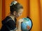 girl in a black dress with gathered blond hair against a background of orange curtains is looking at the globe