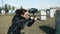 The girl in black shoots from a paintball marker on the open-air ground with gray figures. Side view