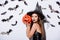 Girl in black witch Halloween costume holding carved spooky pumpkin near white wall