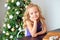 A girl with blond curly hair sits at a table, in the background a Christmas tree, dreams of gifts. Christmas concept
