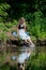 A girl with blond hair on the lake shore has her feet in the water and is swinging them creating a spray of water.