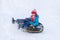 The girl in blue clothes with red hat and scarf descending from the hill covered with a snow on the black rubber ring instead of s