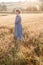 Girl in blue dress standing at dawn in field of ears in golden light of sun. hill and country landscape in background