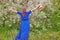 A girl in a blue dress stands with her back against the background of spring nature. Woman and raises hands up near a flowering