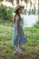 A girl in blue dress and straw hat stands on the shore of the lake