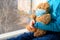 A girl in a blue sweater sits on a windowsill and holds a teddy bear in a medical protective mask.