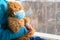 A girl in a blue sweater sits on a windowsill and holds a teddy bear in a medical protective mask.