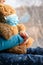 A girl in a blue sweater sits on a windowsill and holds a teddy bear in a medical protective mask.