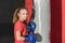 A girl in boxing gloves stands against the background of a sports punching bag in the gym