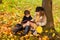 Girl and boy sit under a tree and read a book together in autumn sunny park, children sit in leaves