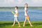 Girl and boy stand on green grass, their backs to each other and play with water pistols, against the blue sky and lake