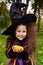 Girl in carnival costume and in hat of witch with little pumpkin in hands on halloween smiling at camera