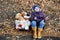 A girl child in a blue hat and yellow boots sits in a pink suitcase with soft toys and looks wistfully to the side. Autumn, dry