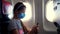 Girl, child in a protective mask uses a mobile, smartphone inside airplane, sitting near illuminator. Resumption of