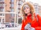 Girl with coffee in warm jacket with fur hood on the street. Winter fashion. Pretty woman in orange hooded jacket in the city.