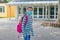 A girl with a crimson, pink backpack stands in front of the school doors in a protective mask. Back to school, starting school