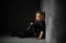 Girl with curly golden hair dressed in black sitting in spotlight on floor close to dark wall, smiling calmly holding hand on knee