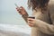 Girl with curly hair in a brown sweater walks on the beach, using cell phone