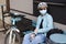 A girl cyclist in a helmet and sunglasses and medical mask is sitting at a table and going to drink water.