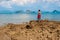 A girl in a dress stands with her back to the camera on a rocky shore near the azure sea. High cliffs in the water on the horizon