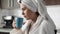 Girl is drinking coffee. Woman in kitchen in white bathrobe with towel on her head sits in front of window and drinks