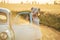 Girl on a dusty country road near a retro car among fields of ripened wheat on a summer day.