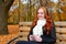 Girl in fall season listen music on audio player with headphones, sit on bench in city park, yellow trees and fallen leaves