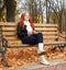 Girl in fall season listen music on audio player with headphones, sit on bench in city park, yellow trees and fallen leaves