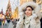 Girl in fur hood walking in Christmas market decorated with holiday lights in the evening. Christmas tree lights on background.