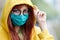 A girl with glasses and a blue reusable medical mask pulls a yellow hood over her head, looks with fear and caution