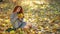 Girl with gorgeous thick curly red hair sits in beam of contour light on blanket of yellow autumn leaves and collects bouquet