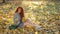 Girl with gorgeous thick curly red hair sits in beam of contour light on blanket of yellow autumn leaves and collects bouquet