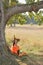 A girl in guise of witch is laughing merrily sitting under huge tree trunk on edge of forest with carved frightening pumpkins