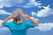 Girl in hat looks at climbing aircraft that flies in clouds on blue sky. White middle aged woman stays back to us and touches her