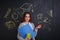 A girl, holding a folder, stationery and a apple, standing next to the board with a picture of science.