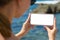 The girl holds horizontally a smartphone with a blank white display on the background of the sea