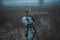 Girl in image of Jeanne d`Arc in armor and with sword in her hands stands on meadow. Back view.